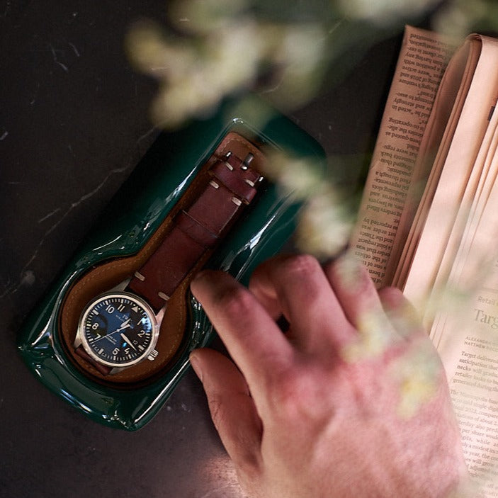 A British Racing Green watch holder with an IWC Pilot's watch in it sitting on a bedside table