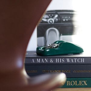 A British Racing Green watch holder with a Rolex Daytona in it sitting on a bookshelf 